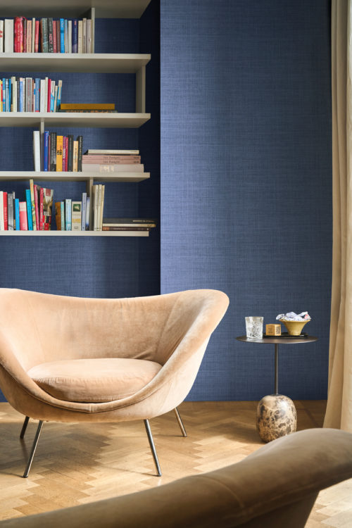 Omexco's High Performance Essence wallpaper in an elegant textured blue, paired with soft velvet seating, open shelving, and a sculptural side table for a sophisticated reading nook.