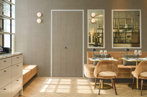 Framing a stylish dining space, Omexco's High Performance Essence wallpaper in a subtle textured taupe pairs with velvet seating, brass accents, and mirrored details for a refined hospitality setting.