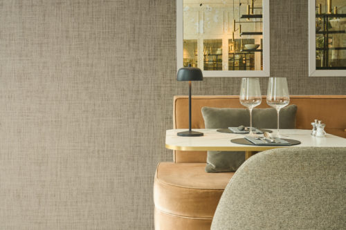 Framing a stylish dining space, Omexco's High Performance Essence wallpaper in a subtle textured taupe pairs with velvet seating, brass accents, and mirrored details for a refined hospitality setting.