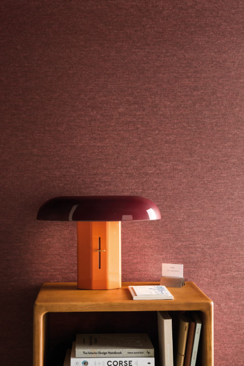 Detail of Omexco's High Performance Essence wallpaper in a rich textured red, styled with a modern sculptural lamp and wooden shelving for a warm, contemporary aesthetic.
