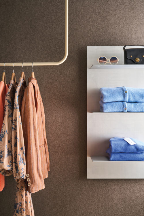 Set against Omexco's High Performance Essence wallpaper in a rich textured brown, a boutique display features patterned garments, soft knitwear, and refined accessories.