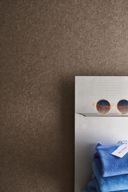 Set against Omexco's High Performance Essence wallpaper in a rich textured brown, a boutique display features patterned garments, soft knitwear, and refined accessories.