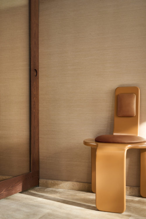 Raumgestaltung mit der Pianura Wandbekleidung von Omexco, der reinste Ausdruck ihres unverwechselbaren Savoir-faire. Die warmbeige strukturierte Wand wird durch einen skulpturalen ockerfarbenen Stuhl mit Lederdetails und eine hölzerne Schiebetür mit Riffelglas ergänzt.