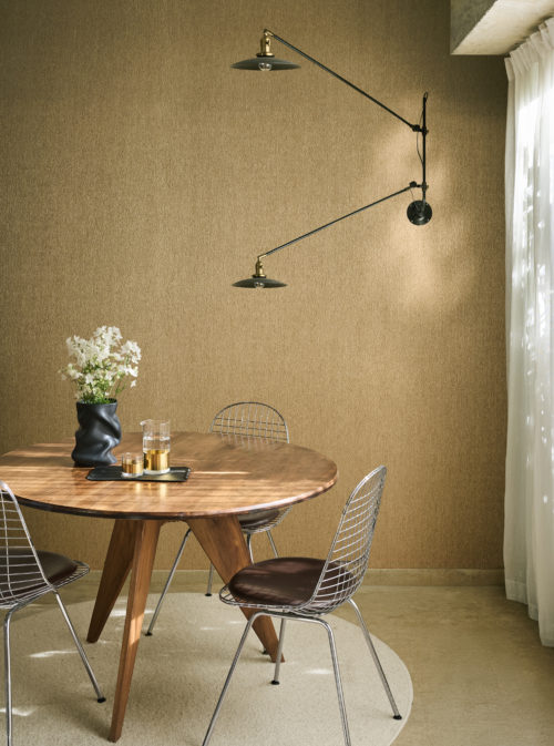 Salle à manger présentant le revêtement mural Pianura d'Omexco dans une teinte dorée chaleureuse, une composition évoquant la délicatesse d'une plume. La scène comprend une table à manger ronde en bois, des chaises en fil de fer et une applique murale noire articulée.