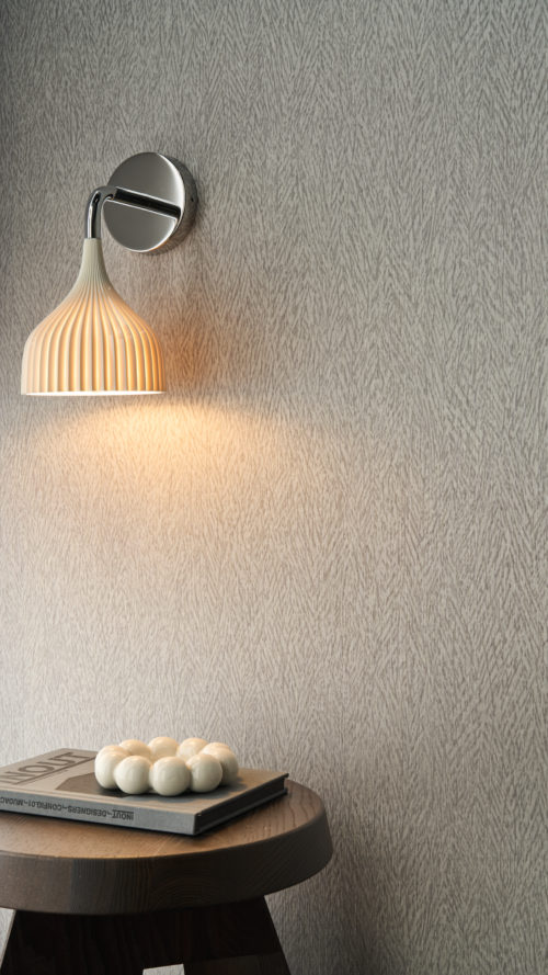 e rapprochée du revêtement mural Pianura d'Omexco, une composition évoquant la délicatesse d'une plume. Le mur gris aux motifs de plumes est éclairé par une applique murale blanche plissée moderne au-dessus d'une table d'appoint ronde en bois.