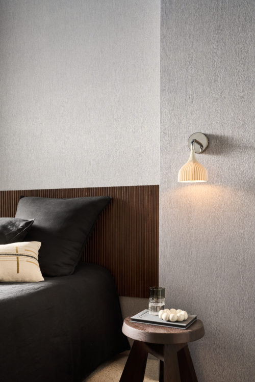Chambre à coucher présentant le revêtement mural gris Pianura d'Omexco, une composition évoquant la délicatesse d'une plume. Une applique murale blanche plissée éclaire le mur texturé au-dessus d'une table de chevet ronde en bois, située à côté d'un lit avec une tête de lit en lattes de bois foncé.