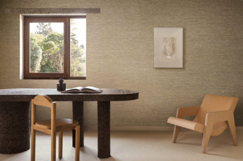 Intérieur serein mettant en valeur le revêtement mural Pianura d'Omexco, fait de très fins fils textiles, réalisés à partir de matières nobles telles que le lin. Les murs texturés chaleureux servent de toile de fond à une table d'aspect pierre, des chaises en bois et une petite fenêtre encadrant de la verdure.