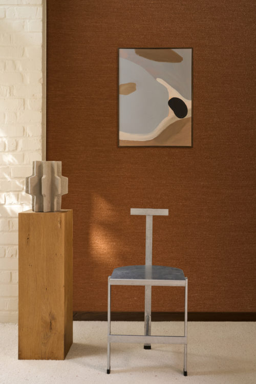 Espace intérieur exposant le revêtement mural Pianura d'Omexco dans un riche ton brun cuivré, composé d'un fil de laine duveteux, comme effleuré par le vent. Une peinture abstraite est accrochée au mur texturé, accompagnée d'une chaise en métal minimaliste et d'une plinthe en bois massif exposant une sculpture grise.