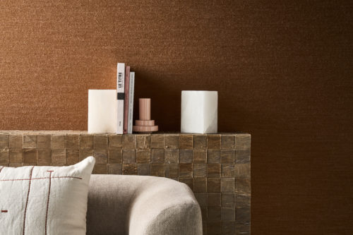 Interieurdetail mit der Pianura Wandbekleidung von Omexco in einem rostbraunen Farbton aus weicher, luftiger Wolle, die an sanft vom Wind bewegte Fasern erinnert. Die stark strukturierte Wand dient als Hintergrund für einen massiven Konsolentisch mit Büchern und Marmorbuchstützen hinter einem hellen Sofa.