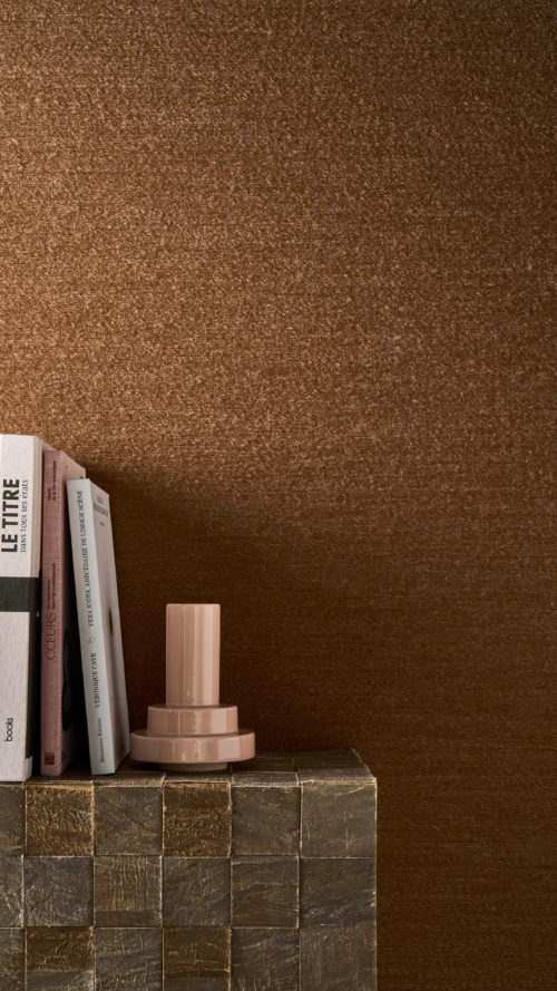 Détail en gros plan du revêtement mural Pianura d'Omexco dans une teinte brun rouille, composé d'un fil de laine duveteux, comme effleuré par le vent. Le mur texturé sert de toile de fond à une pile de livres et à un vase rose brillant moderne posés sur un bloc texturé.