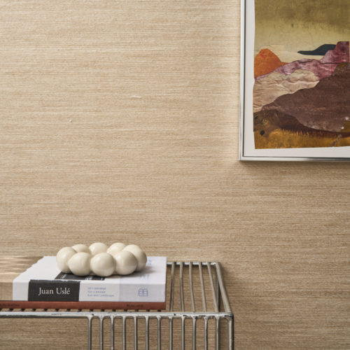 Interior detail showcasing the light beige Pianura wallcovering by Omexco, made of soft, airy wool, as if gently stirred by the wind. The textured wall is decorated with a framed abstract landscape artwork and sits behind a wire table holding a book and a bubble-shaped decorative object.