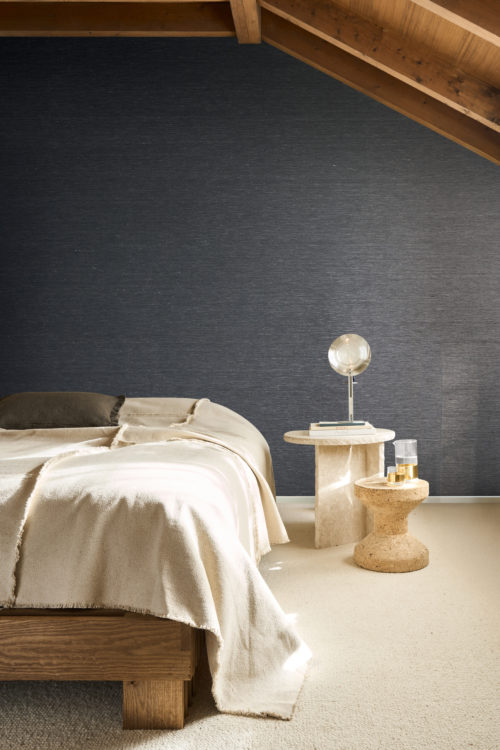 Intérieur de chambre à coucher mettant en valeur le revêtement mural gris foncé Pianura d'Omexco, fait de fils textiles délicats en lin torsadé et bicolore, aux nuances subtiles. L'élégant mur texturé est positionné derrière un lit recouvert d'une couverture claire texturée et une table d'appoint sculpturale en pierre avec une lampe ronde.