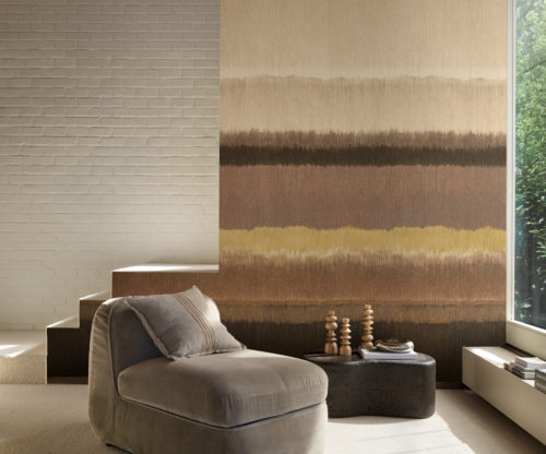 Un intérieur spacieux présentant le revêtement mural Pianura à rayures horizontales d'Omexco, une évocation de paysages embrumés, animée par de riches nuances de couleur. Le design s'écoule avec des bandes terreuses de brun, de beige et de moutarde à côté d'un mur de briques peintes en blanc, encadrant un espace confortable avec un fauteuil beige.