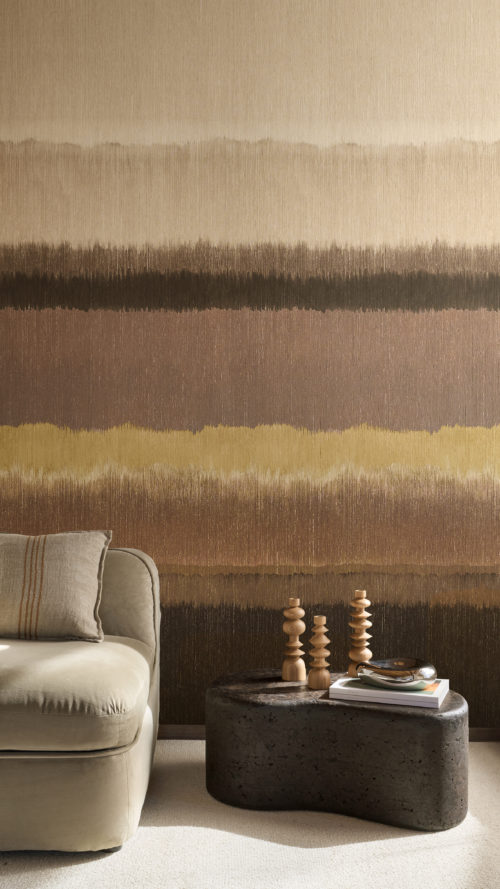Intérieur exposant le revêtement mural Pianura à rayures horizontales d'Omexco, offrant une évocation de paysages embrumés, animée par de riches nuances de couleur. Les bandes superposées de brun, beige et moutarde créent une atmosphère chaleureuse derrière un canapé beige clair et une table sculpturale noire avec des bougeoirs en bois.