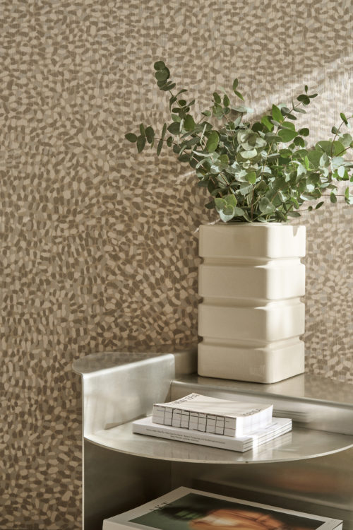 Gros plan du revêtement mural Pianura beige clair et gris d'Omexco, affichant des accents de pelage, qui anime les murs avec caractère. Le délicat motif tacheté forme une toile de fond pour un vase blanc carré nervuré contenant des branches d'eucalyptus vert sur une table métallique.