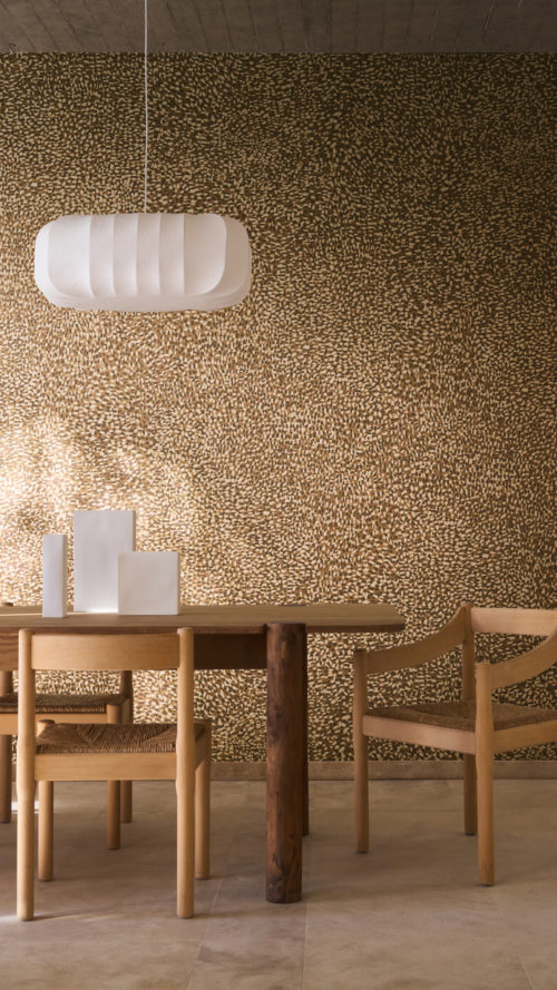 Dining area featuring the patterned Pianura wallcovering by Omexco with echoes of animal coats, bringing walls to life with instinct and movement. The vibrant brown and white speckled wall sets a lively backdrop for a wooden dining table, chairs with woven seats, and a white paper pendant lamp.