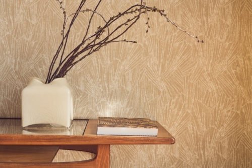 Nahaufnahme der goldbeigen Whisper Wandbekleidung von Omexco mit einem stilisierten Motiv, inspiriert von feinen mineralischen Strukturen und Muscheln. Die wunderschön strukturierte Wand befindet sich hinter einem Holztisch im Mid-Century-Stil mit einer weißen Vase und dekorativen Zweigen.