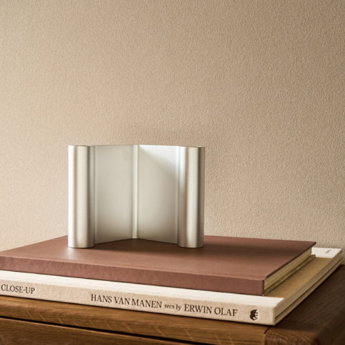 Close-up view of the beige Whisper wallcovering by Omexco, showing delicately dusted rice husk powder tracing a fine pattern of mineral veins. The organically textured wall acts as a minimalist backdrop for a curved silver metallic object resting on a stack of books