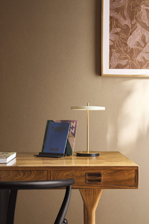 A home office setup featuring the brown Whisper wallcovering by Omexco, where delicately dusted rice husk powder traces a fine pattern of mineral veins. The warmly textured wall complements a wooden desk, a modern brass lamp, and a framed abstract artwork.
