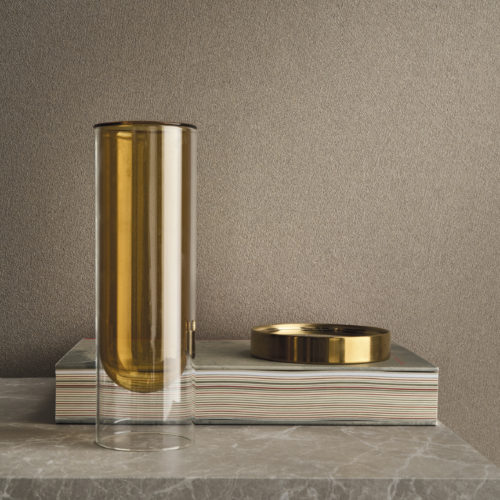 EN: Detail of the warm brown Whisper wallcovering by Omexco, where delicately dusted rice husk powder traces a fine pattern of mineral veins. The subtly textured wall provides a rich background for a tall amber glass vase and a golden dish resting on stacked books.