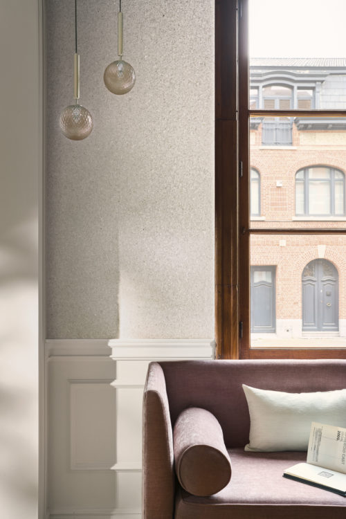 Ein eleganter Wohnraum mit der hellen Whisper Wandbekleidung von Omexco, versehen mit zarten Muschelfragmenten, die einen sanften, perlmuttartigen Schimmer erzeugen. Die funkelnde Wand befindet sich über einer klassischen weißen Täfelung, neben einem Fenster und einem pflaumenfarbenen Sofa, das von bernsteinfarbenen Glaspendelleuchten beleuchtet wird.