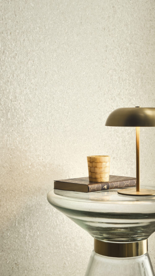 Interior detail highlighting the sparkling white Whisper wallcovering by Omexco, beautifully textured with finely applied shell fragments lending a soft, pearlescent warmth. The luminous wall sets a serene tone behind a sculptural glass table adorned with a modern brass lamp and a mosaic candle holder.