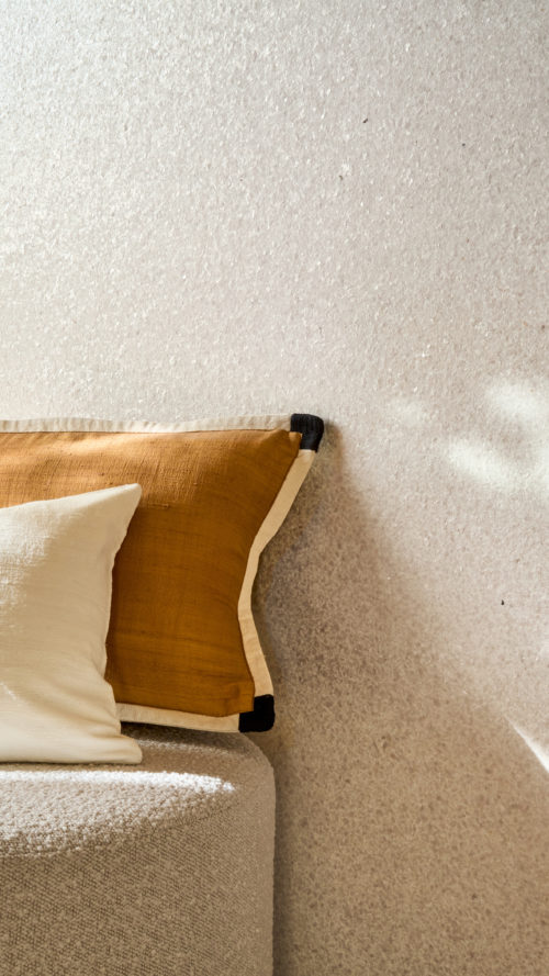 Detailaufnahme der hellen Whisper Wandbekleidung von Omexco, verziert mit zarten Muschelfragmenten, die einen sanften, perlmuttartigen Schimmer erzeugen. Die leuchtend strukturierte Wand wird von weichem Sonnenlicht erhellt und ergänzt einen strukturierten Pouf mit weißen und ockerfarbenen Kissen im Vordergrund.