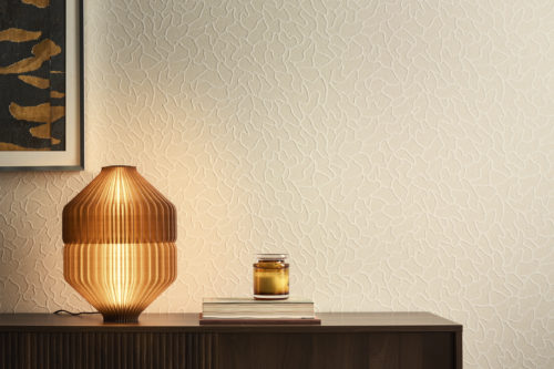 Ein stilvolles Interieur mit der hellen Whisper Wandbekleidung von Omexco, ein organisches Motiv, dessen lebendige Struktur durch sanft verteiltes Marmorpuder auf einem Mica-Hintergrund entsteht. Die dezent gemusterte Wand ergänzt ein Holz-Sideboard mit einer leuchtenden bernsteinfarbenen gerippten Tischlampe.