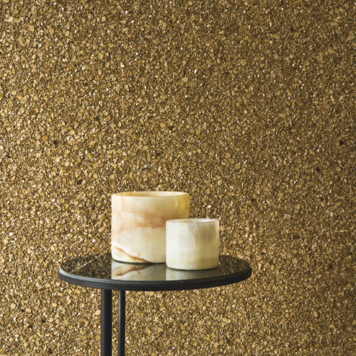 Nahaufnahme der strahlend goldenen Whisper Wandbekleidung von Omexco aus strukturiertem Mica in einer neuen Farbpalette. Die reich besetzte Wand dient als luxuriöser Hintergrund für zwei Alabastergefäße auf einem kleinen schwarzen Tisch.