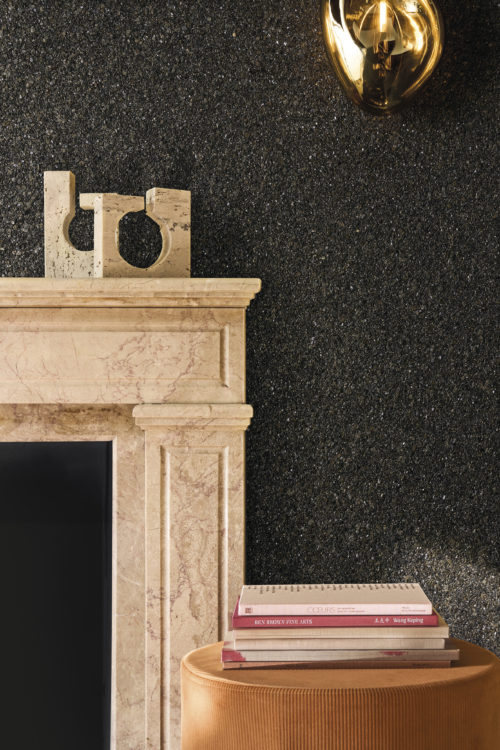 Eleganter Raum mit der dunklen Whisper Wandbekleidung von Omexco aus strukturiertem Mica in einer neuen Farbpalette. Die funkelnde schwarze Wand bildet einen schönen Kontrast zu einem klassischen Kamin aus rosafarbenem Marmor und einer modernen bernsteinfarbenen Wandleuchte.