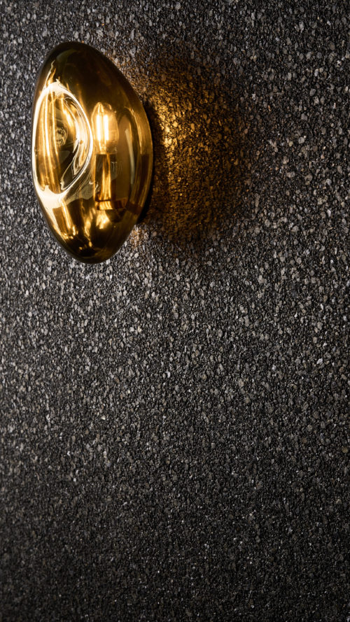 Detail der dunklen Whisper Wandbekleidung von Omexco aus strukturiertem Mica in einer neuen Farbpalette. Die tief strukturierte, funkelnd schwarze Wand wird von einer bernsteinfarbenen Glaswandleuchte dramatisch erhellt.