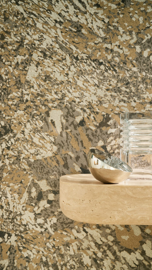Detailaufnahme der gemusterten Whisper Wandbekleidung von Omexco, die einen kontrastreichen Marmor mit malerischen Akzenten zeigt, veredelt durch einen sanft irisierenden Hintergrund. Eine beige Steinkonsole im Vordergrund präsentiert eine kleine silberne Schale und gerippte Glasobjekte.