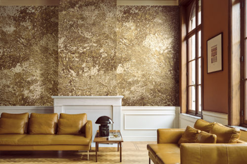 Elegantes Wohnzimmer mit der goldenen Whisper Wandbekleidung von Omexco, ein kontrastreicher Marmor mit malerischen Akzenten, veredelt durch einen sanft irisierenden Hintergrund. Die majestätische Wand umgibt einen klassischen weißen Kamin, ergänzt durch warme gelbe Ledersofas und einen Parkettboden.
