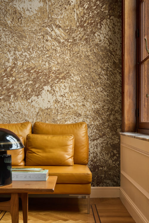 Nahaufnahme eines Wohnraums, dekoriert mit der Whisper Wandbekleidung von Omexco, ein kontrastreicher Marmor mit malerischen Akzenten, veredelt durch einen sanft irisierenden Hintergrund. Die satte, goldbraun gemusterte Wand ergänzt perfekt ein warmes gelbes Ledersofa und eine dunkle, glänzende Tischlampe.