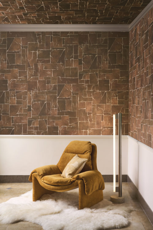 A striking interior featuring the brown Whisper wallcovering by Omexco on both the walls and ceiling, presenting a marble-effect marquetry with finely inlaid detailing on a mica background. The room includes a mustard yellow armchair on a white fluffy rug and a modern tubular floor lamp
