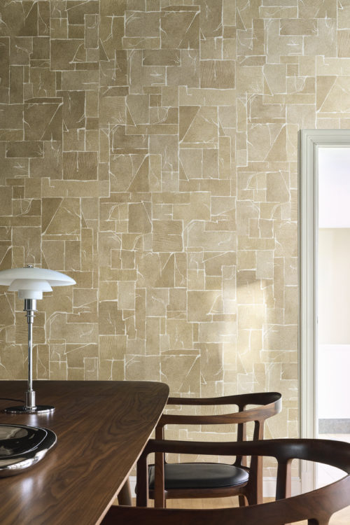 Raumgestaltung mit der goldenen Whisper Wandbekleidung von Omexco, die Intarsien in Marmoroptik mit fein ausgearbeiteten Details auf einem Mica-Hintergrund zeigt. Die gemusterte Wand bietet einen warmen Hintergrund für einen dunklen Holztisch, gebogene Holzstühle und eine klassische weiße Tischlampe.