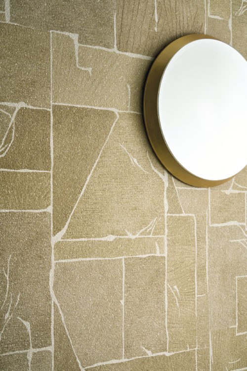 Detail of the golden Whisper wallcovering by Omexco, showcasing a marble-effect marquetry with finely inlaid detailing on a mica background. The geometric, textured wall is gracefully adorned with a simple round mirror with a brass frame.