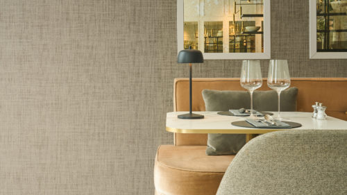 Framing a stylish dining space, Omexco's High Performance Essence wallpaper in a subtle textured taupe pairs with velvet seating, brass accents, and mirrored details for a refined hospitality setting.