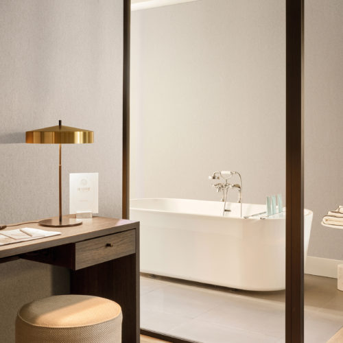 Softly textured in beige, Omexco's High Performance Essence wallpaper sets a tranquil tone, paired with a dark wood desk, golden lamp, and a glimpse of a spa-like bathroom with freestanding tub.
