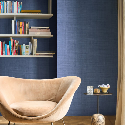 Omexco's High Performance Essence wallpaper in an elegant textured blue, paired with soft velvet seating, open shelving, and a sculptural side table for a sophisticated reading nook.
