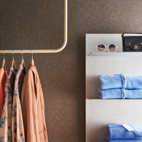 Set against Omexco's High Performance Essence wallpaper in a rich textured brown, a boutique display features patterned garments, soft knitwear, and refined accessories.