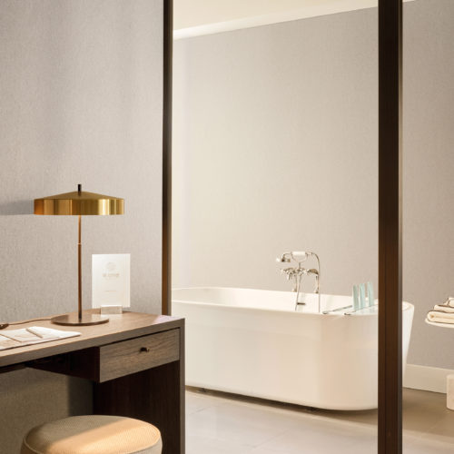 Softly textured in beige, Omexco's High Performance Essence wallpaper sets a tranquil tone, paired with a dark wood desk, golden lamp, and a glimpse of a spa-like bathroom with freestanding tub.