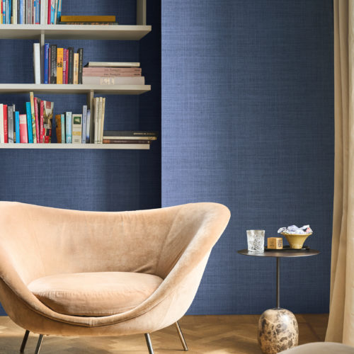 Omexco's High Performance Essence wallpaper in an elegant textured blue, paired with soft velvet seating, open shelving, and a sculptural side table for a sophisticated reading nook.