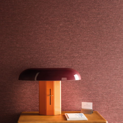 Detail of Omexco's High Performance Essence wallpaper in a rich textured red, styled with a modern sculptural lamp and wooden shelving for a warm, contemporary aesthetic.