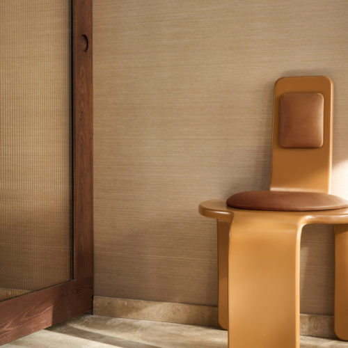 Intérieur présentant le revêtement mural Pianura d'Omexco, expression de leur savoir-faire emblématique. Le mur texturé beige chaud est mis en valeur par une chaise sculpturale ocre avec des détails en cuir et une porte coulissante en bois avec verre cannelé.