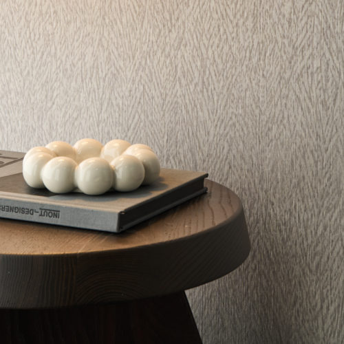 e rapprochée du revêtement mural Pianura d'Omexco, une composition évoquant la délicatesse d'une plume. Un mur gris aux motifs de plumes en une table d'appoint ronde en bois.