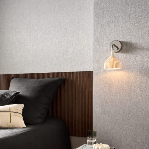 Bedroom setting featuring the grey Pianura wallcovering by Omexco, a delicate composition, imbued with the protective nature of plumage. A white pleated wall lamp illuminates the textured wall above a round wooden bedside table, situated next to a bed with a dark slatted wooden headboard.