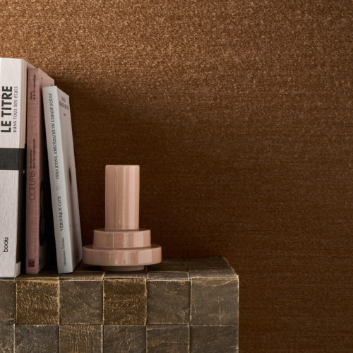Détail en gros plan du revêtement mural Pianura d'Omexco dans une teinte brun rouille, composé d'un fil de laine duveteux, comme effleuré par le vent. Le mur texturé sert de toile de fond à une pile de livres et à un vase rose brillant moderne posés sur un bloc texturé.