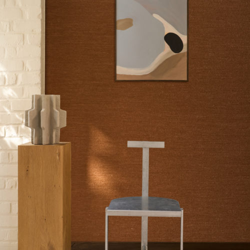 Interior space displaying the Pianura wallcovering by Omexco in a rich copper-brown tone, made of soft, airy wool, as if gently stirred by the wind. An abstract painting hangs on the textured wall, accompanied by a minimalist metal chair and a solid wooden plinth displaying a grey sculpture.