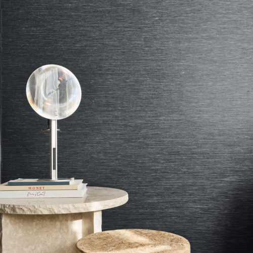 Vue rapprochée du revêtement mural gris foncé Pianura d'Omexco, fait de fils textiles délicats en lin torsadé et bicolore, aux nuances subtiles. Le mur finement texturé offre une toile de fond sombre et élégante pour une lampe de table ronde translucide reposant sur des livres empilés sur une table en pierre claire.